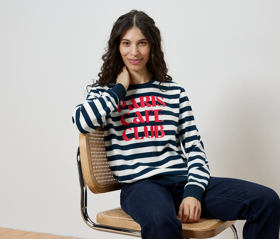 Frau sitzt auf einem Stuhl und trägt ein gestreiftes Sweatshirt mit der Aufschrift "Paris Cafe Club" und Jeans.