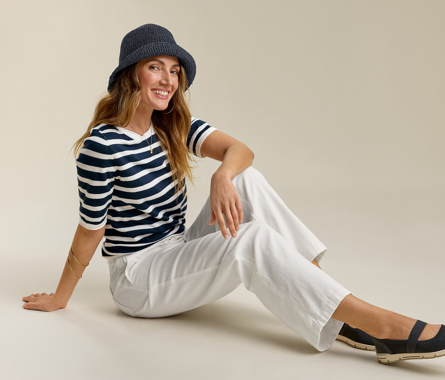Frau sitzt mit Wide-Komfort-Jeans, gestreiftem Shirt und Hut. Sie lächelt.