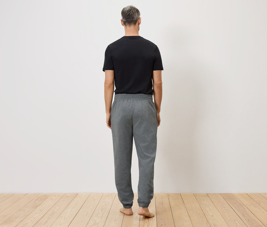 Mann von hinten in schwarzem T-Shirt und grauer Jogginghose vor weißer Wand und Holzboden.