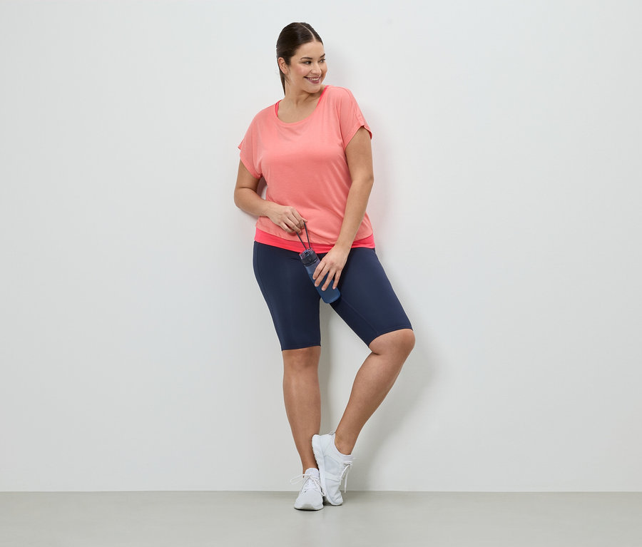 Frau in Sportbekleidung mit Trinkflasche lehnt an Wand. Sie trägt ein korallfarbenes T-Shirt, blaue Shorts und weiße Turnschuhe.
