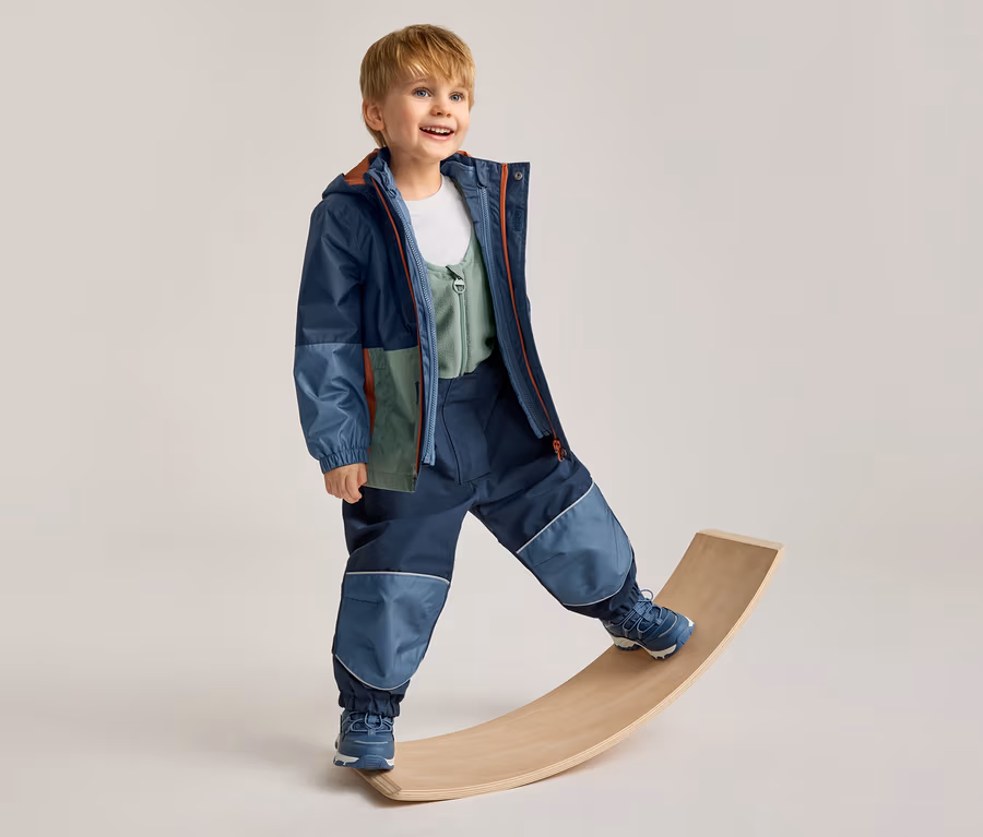 Lächelnder Junge in einer mehrfarbigen Jacke und dunkelblauen Hose mit hellblauen Patches, der auf einem Holzbrett balanciert.