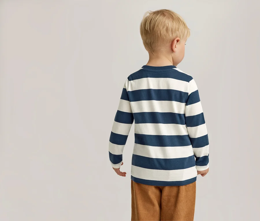 Rückansicht eines Kindes mit blonden Haaren in einem blau-weiß gestreiften Langarmshirt und brauner Cordhose vor einem hellen Hintergrund.