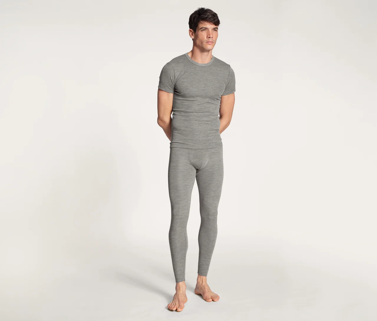Ein Mann steht barfuß vor einem weißen Hintergrund und trägt ein graues kurzärmeliges T-Shirt und eine passende lange Unterhose. Seine Hände sind hinter seinem Rücken verschränkt.