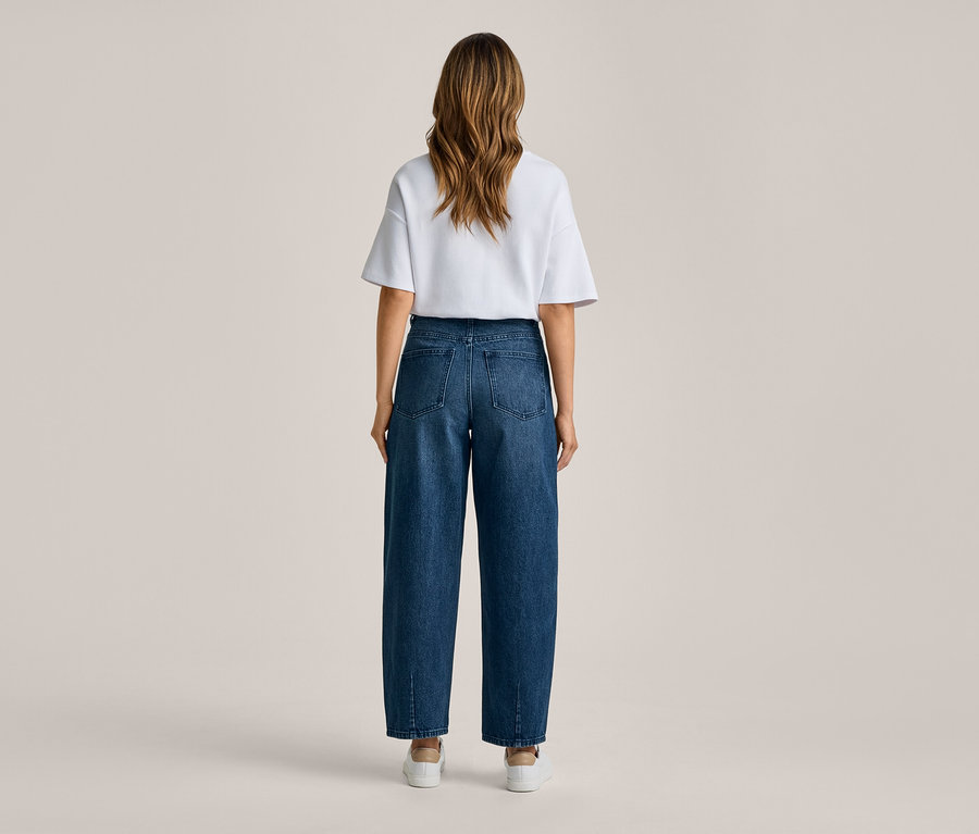 Frau steht mit dem Rücken zum Betrachter, trägt ein weißes T-Shirt, dunkelblaue weite Jeans und weiße Sneaker.