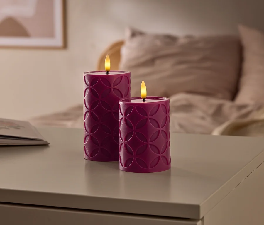 Zwei lila LED-Kerzen mit geometrischem Muster leuchten auf einem Tisch, daneben ein offenes Buch und ein Bett im unscharfen Hintergrund.