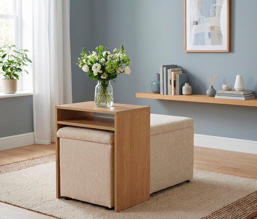 Ein hellbrauner Beistelltisch mit zwei beigen Hockern, von denen einer unter dem Tisch verstaut ist, steht auf einem hellen Teppich. Auf dem Tisch steht eine Vase mit weißen und grünen Blumen. Im Hintergrund sind eine hellblaue Wand, ein Fenster mit weißen Vorhängen und ein Regal mit Büchern und Dekoration zu sehen.