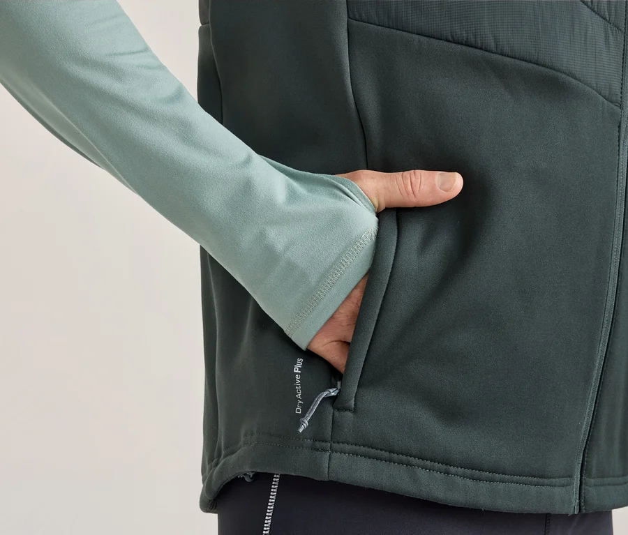 Person trägt hellgrünes Langarmshirt mit Daumenlöchern und dunkelgraue Weste, die Hand steckt in der Westentasche. Auf der Weste steht „Dry Active Plus“.