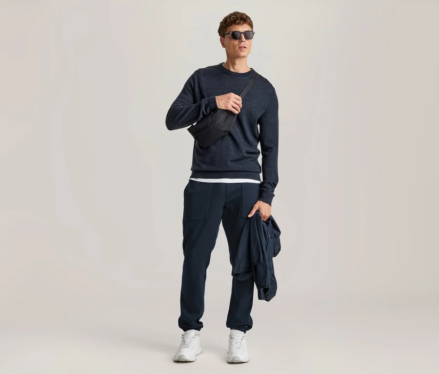 Mann in einem dunkelblauen Pullover, dunkelblauer Hose, weißen Turnschuhen und Sonnenbrille, mit einer schwarzen Bauchtasche über der Brust und einer dunkelblauen Jacke in der Hand.