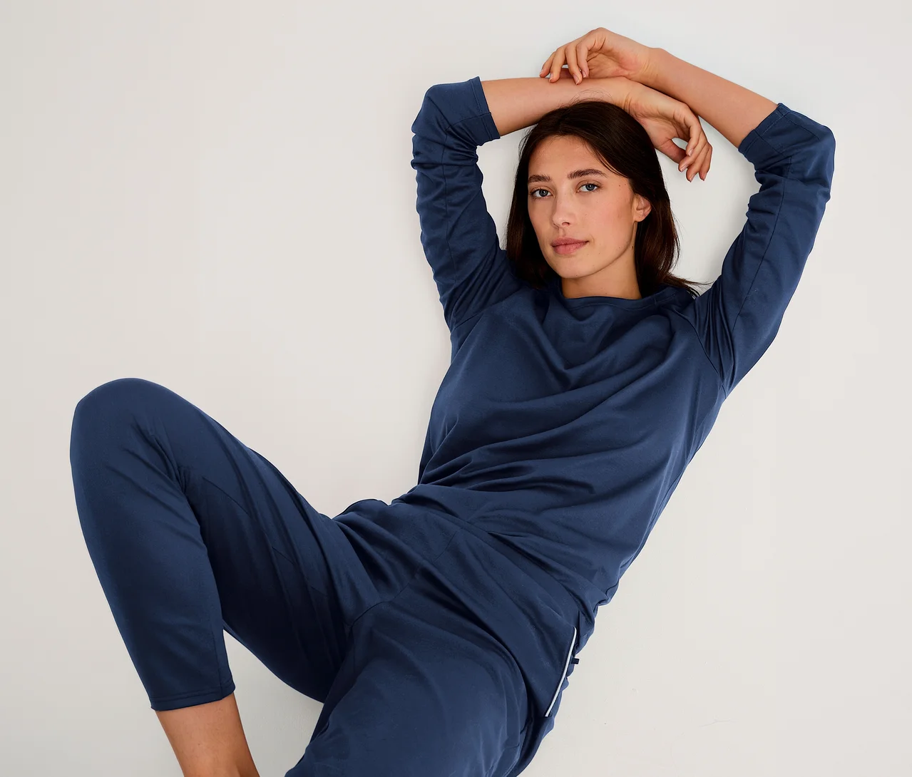 Frau in marineblauem Langarmshirt und passender Hose, die mit erhobenen Armen an einer weißen Wand lehnt und in die Kamera blickt.