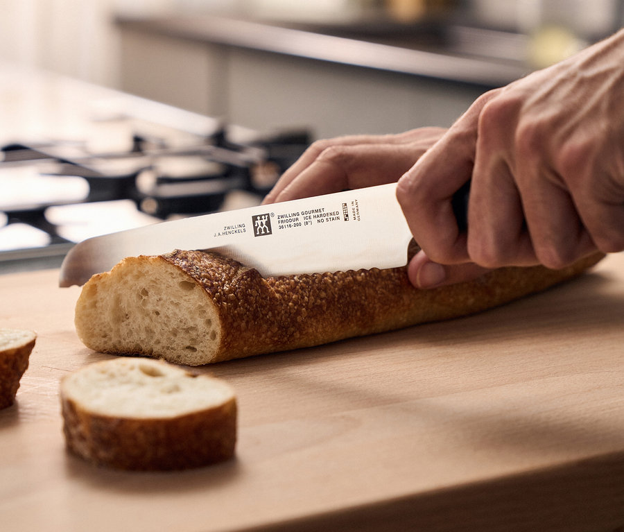 Hände schneiden ein Baguette auf einem Holzschneidebrett mit einem ZWILLING Messer, zwei Scheiben sind bereits geschnitten.