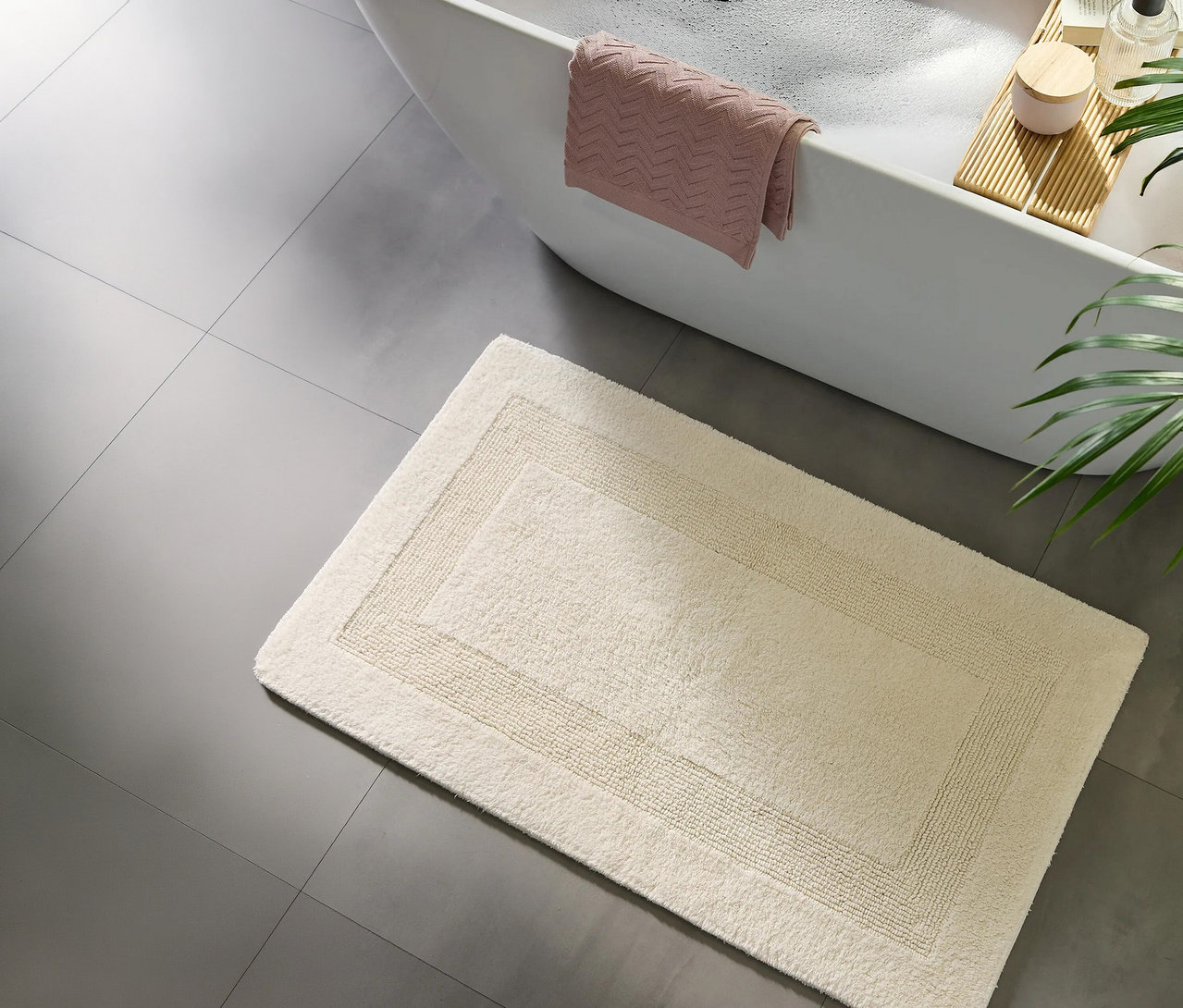 Blick von oben auf ein Badezimmer mit einer weißen Badewanne voller Schaum, einem rosa Handtuch über dem Rand und einem hölzernen Badewannenbrett. Eine cremefarbene, rechteckige Badematte mit dezentem Rand liegt auf dem grauen Fliesenboden vor der Wanne.