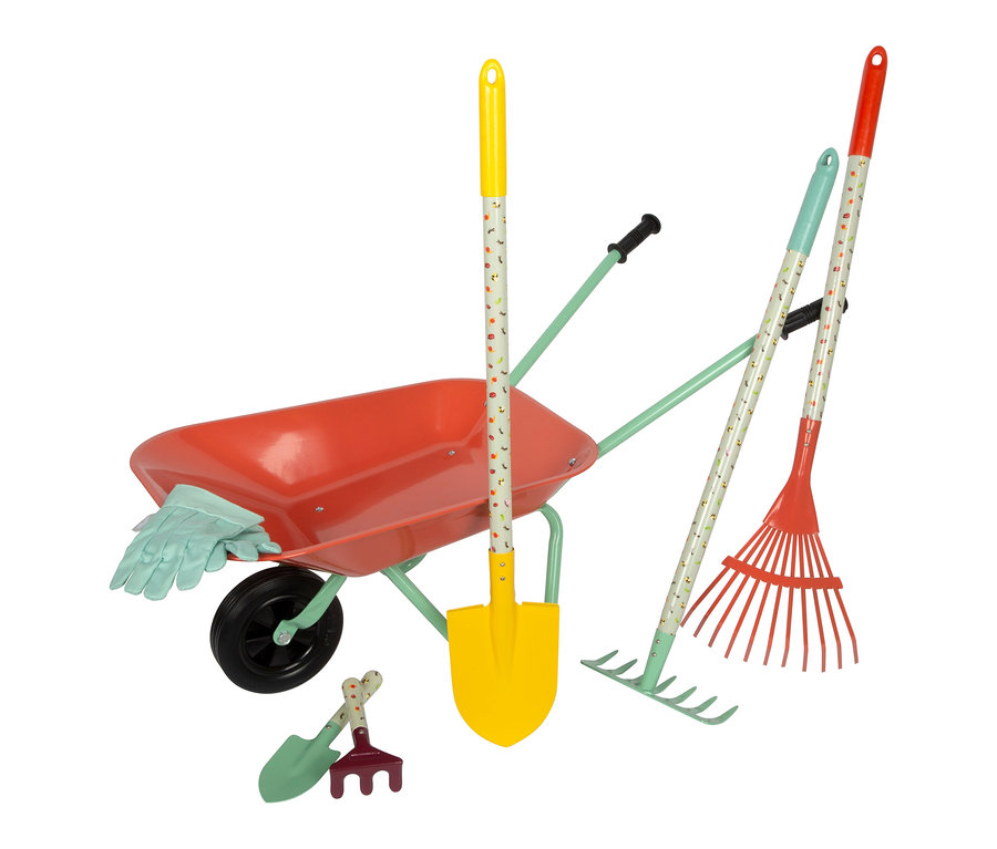 Eine rote small foot Schubkarre mit hellgrünem Gestell, gefüllt mit hellblauen Handschuhen, steht neben einer gelben Schaufel, zwei Rechen und kleinem Gartenwerkzeug.