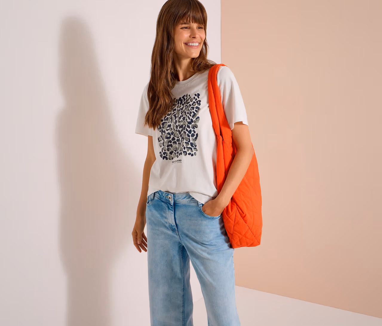 Lächelnde Frau mit langen braunen Haaren trägt ein weißes CECIL Jerseyshirt mit Leopardenmuster, hellblaue Jeans und eine orangefarbene gesteppte Tragetasche über der Schulter.