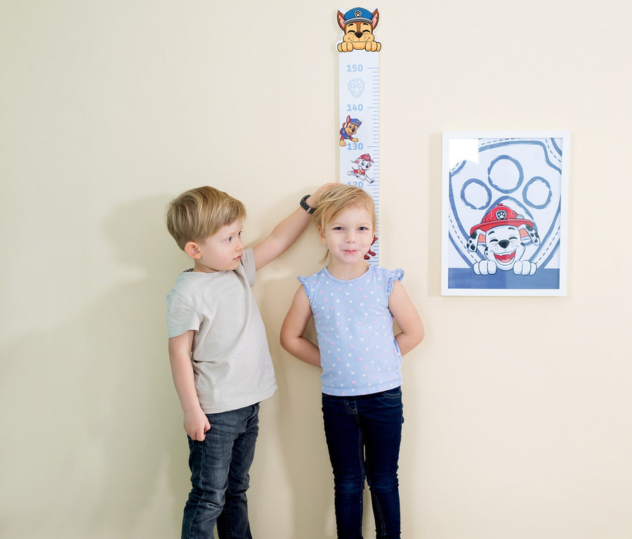 Ein Junge misst die Größe eines Mädchens an einem Paw Patrol-Messlatte, neben der ein gerahmtes Paw Patrol-Bild hängt.