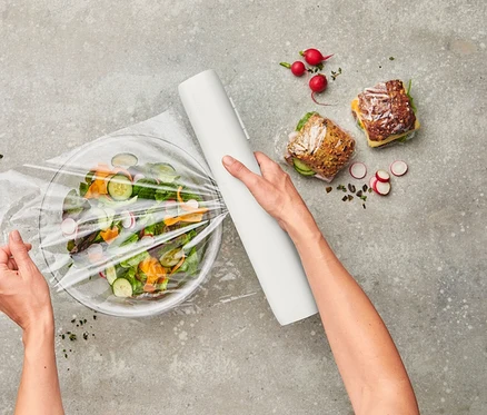 Draufsicht: Person verpackt Salat in Schüssel mit Frischhaltefolie. Daneben: Dressing, belegte Brote und Radieschen.