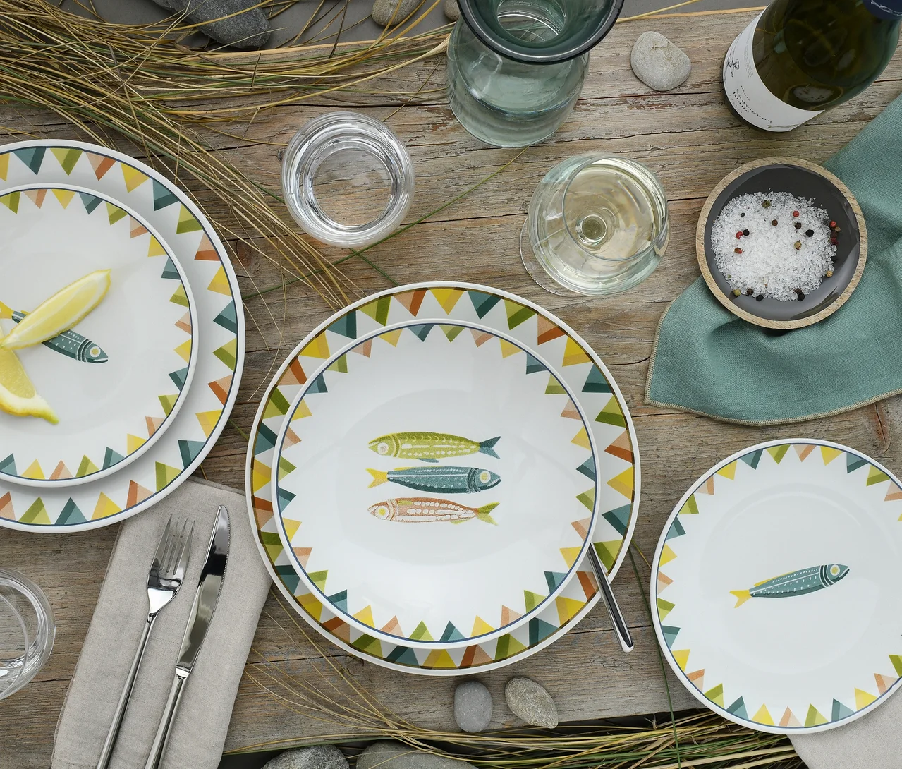 Draufsicht auf einen rustikalen Holztisch, gedeckt mit drei weißen creaTable „Sardinhas Pastell“ Tellern, die bunte Sardinenillustrationen und einen Rand aus grünen, gelben und braunen Dreiecken zeigen. Zwei Teller zeigen eine einzelne Sardine, einer ein Trio. Ebenfalls auf dem Tisch sind Besteck, Gläser, eine Weinflasche, Salz und eine grüne Serviette, umgeben von trockenem Gras und Kieselsteinen.