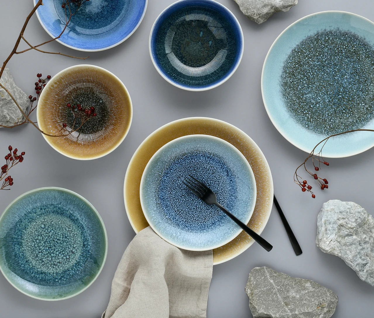 Verschiedene creaTable Caldera Schalen und Teller in Blau, Grün und Braun mit gesprenkelten Texturen sind auf einer grauen Oberfläche angeordnet. Eine schwarze Gabel und ein Messer liegen auf einem blau gesprenkelten Teller, der auf einem größeren gelb-braunen Teller liegt, neben einer Leinen-Serviette. Dekorative Zweige mit roten Beeren und graue Steine sind ebenfalls sichtbar.